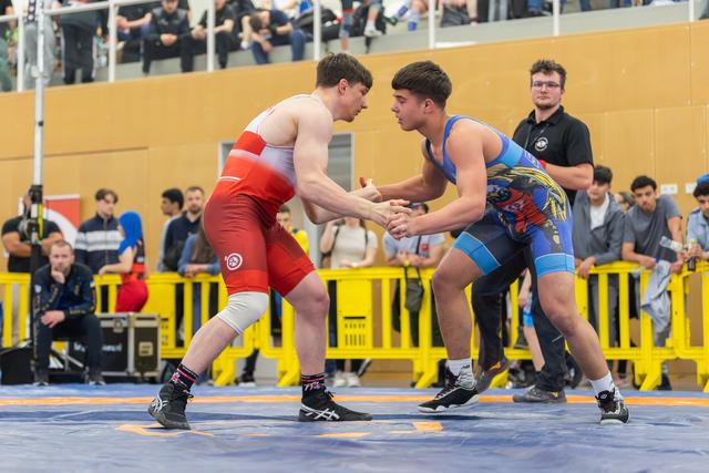 Two young wrestlers lock hands in an intense grip battle, focused and poised on a blue mat before a crowd.