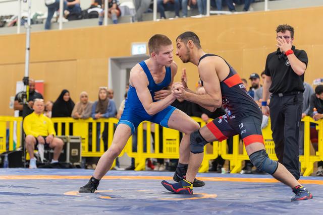 Two young wrestlers lock hands in tense standoff on blue mat, both crouched low, focused and determined.