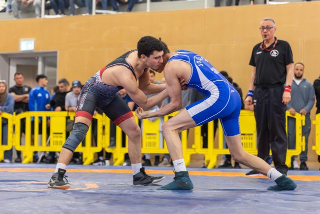 Two young wrestlers in focused stance, grappling on blue mat at indoor sports hall, referee watching nearby.