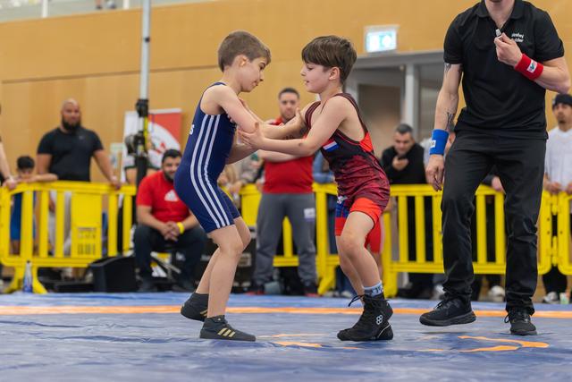 Two focused young boys in singlets grip each other's arms on a blue mat, referee watching closely nearby.