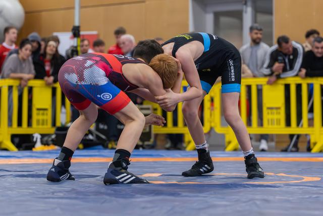 Two young wrestlers locked in a crouched stance on the mat, focused and tense, with a crowd watching behind barriers.