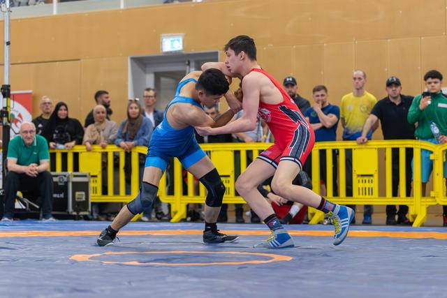 Two young wrestlers clash on the mat—blue uniform lunges forward aggressively against red, crowd watching intently behind yellow barriers.
