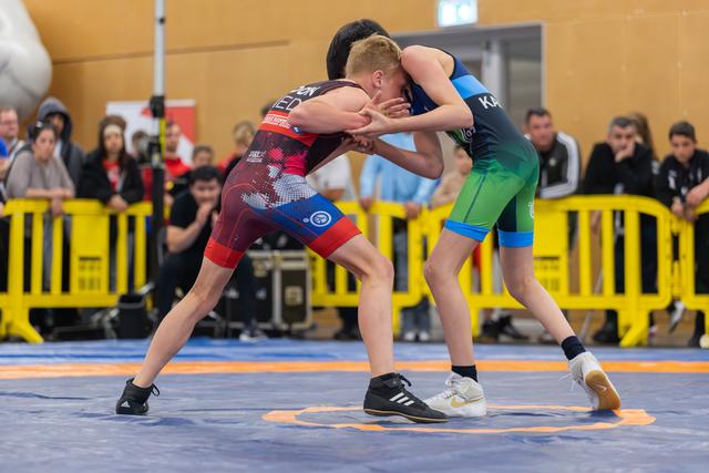 Two young wrestlers lock up in an intense standing clinch on a blue mat, crowd watching from behind yellow barriers.