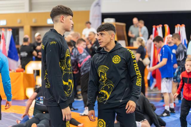 Two teenage wrestlers in black and gold Freiburg tracksuits face each other with focused, determined expressions on the mat.
