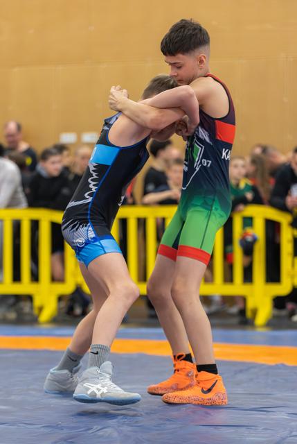 Two young wrestlers lock arms in a standing clinch on a blue mat, both straining with focused intensity during a bout.