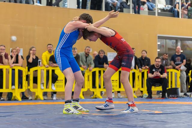 Two young wrestlers lock arms in a tense standoff on a blue mat, watched by a crowd of spectators behind yellow barriers.