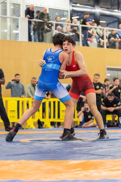 Teen in red singlet grips opponent in blue, both focused and tense on the mat, crowd watching from above.