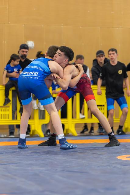 Two teenage wrestlers locked in a tight clinch on a blue mat, both straining forward with intense focus during a bout.