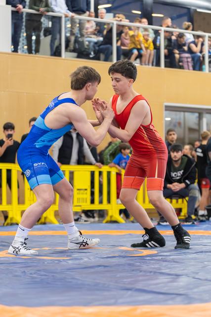 Two young wrestlers in blue and red singlets lock hands in a tense standoff on a blue mat, crowd watching.