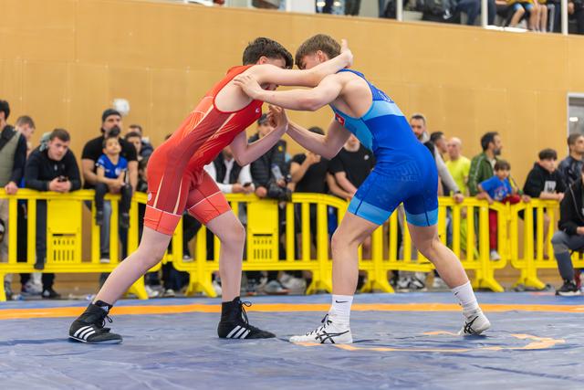 Two young wrestlers lock arms in intense standoff on blue mat, crowd watching behind yellow barriers.