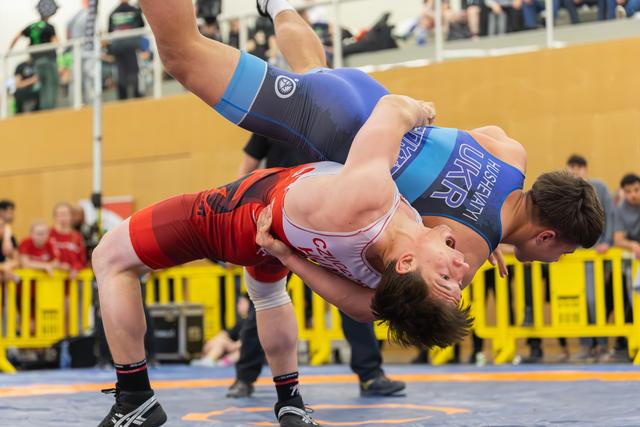 Ukrainian wrestler in blue executes a powerful throw, lifting his red-singlet opponent overhead in an indoor arena.
