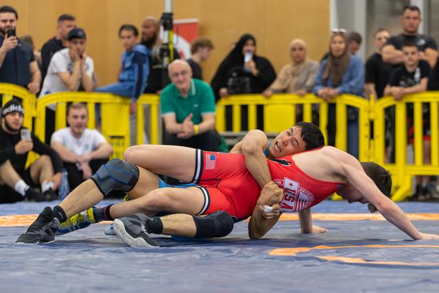 US wrestler grimaces in pain as opponent attempts a pin during an indoor match, crowd watching intently.