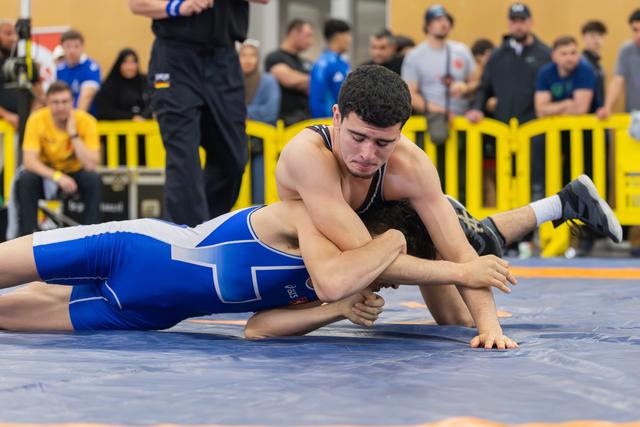 Young wrestler in black singlet dominates opponent in blue, pinning him face-down on the mat with focused intensity.