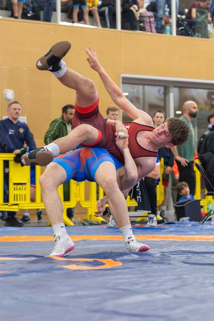 Wrestler in blue singlet lifts and throws opponent in red, mid-air, during an indoor match with spectators watching.