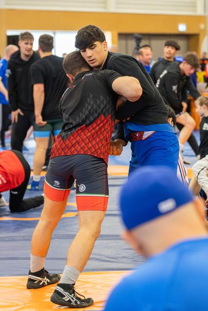 Two wrestlers lock in a tight clinch on an orange mat, surrounded by other athletes training in a busy sports hall.