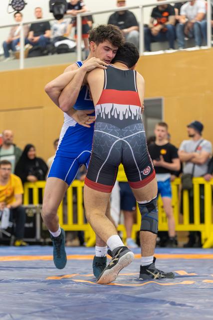 Two wrestlers locked in a tight clinch, one lifted off the mat mid-match, crowd watching in the background.