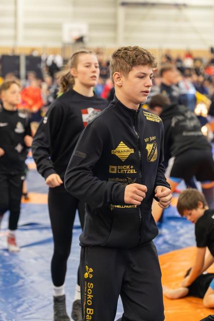 Focused teen wrestler in black sponsor jacket jogs during warm-up in a busy indoor sports hall.