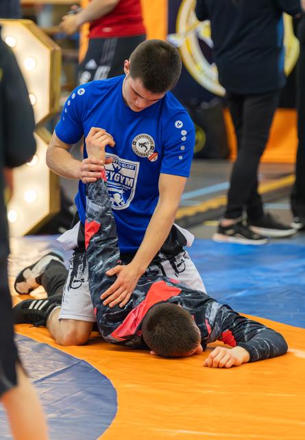 Teen in blue gym shirt applies submission hold on opponent lying face-down on orange mat during match.