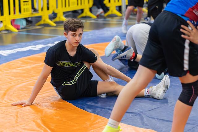 Teen wrestler in black sits on the mat, composed and focused, watching others practice in a busy sports hall.