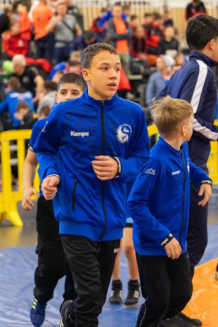Focused teenage boy in blue Kempa jacket leads younger teammates across the mat at an indoor sports event.