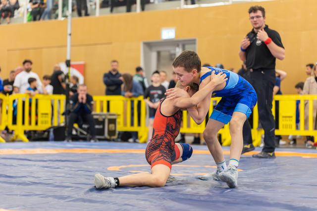 Two young wrestlers locked in a grapple, one in red kneeling, the other in blue pressing forward with focus.