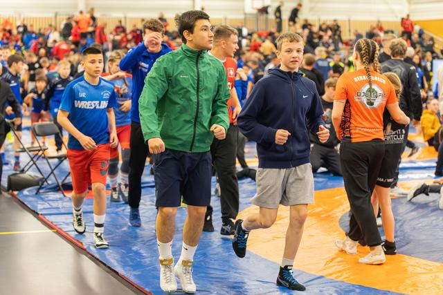 Two teenage boys jog confidently across the mat in a busy sports hall, surrounded by athletes warming up.