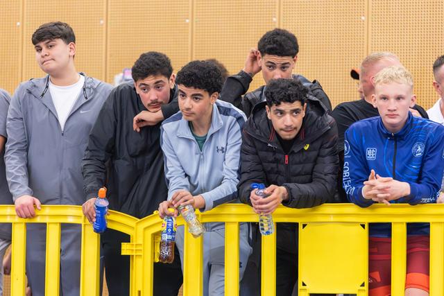 Group of teenage athletes lean on a yellow barrier, watching intently with focused, serious expressions at an indoor sports event.