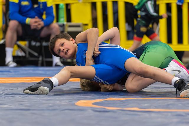 Young boy in blue singlet pins opponent to mat, face showing intense focus during youth wrestling match.