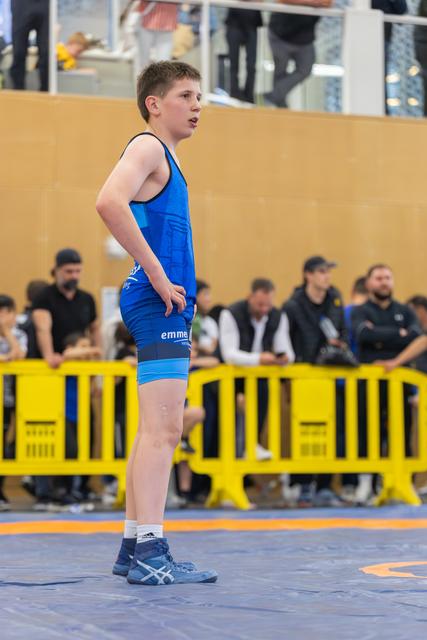 Young wrestler in blue singlet stands alert on the mat, mouth open, hands ready, focused between bouts.