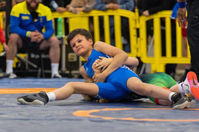 Young boy in blue singlet wrestles opponent to the mat, gripping tightly with fierce concentration during youth bout.