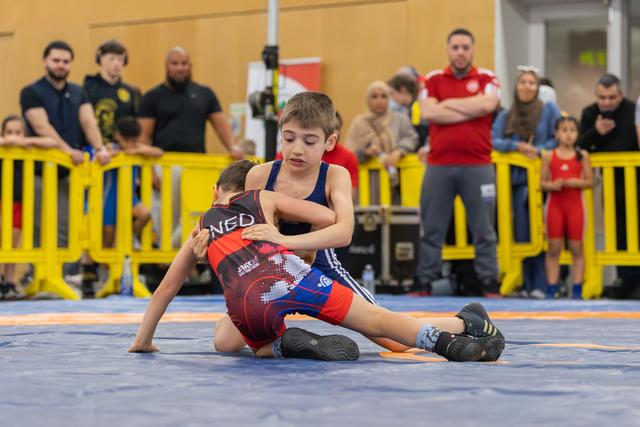 Two young boys wrestle on a blue mat; the boy in blue controls his opponent with a firm grip, focused and determined.