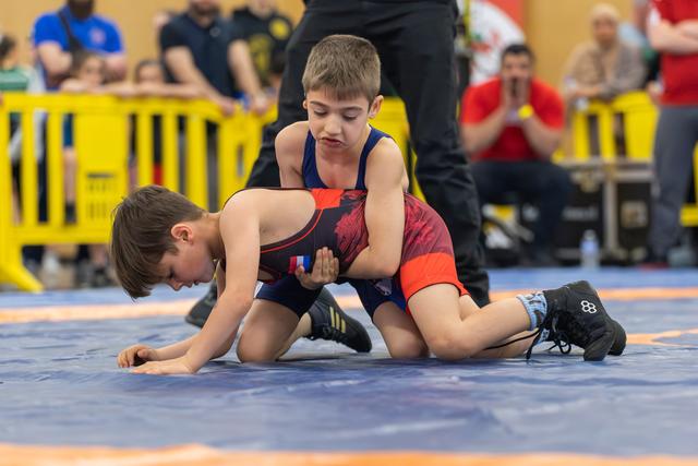 Two boys wrestle on a blue mat; the boy on top focuses intensely, controlling his opponent with a firm grip.