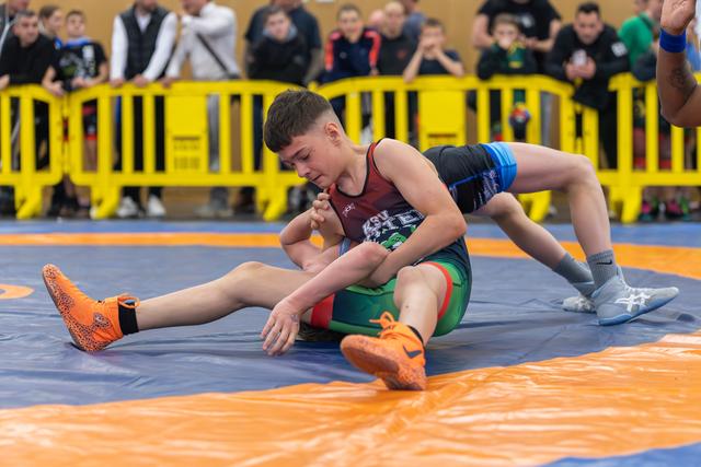 Focused young wrestler controls his opponent on the orange mat, bearing down with intensity before a crowd of spectators.