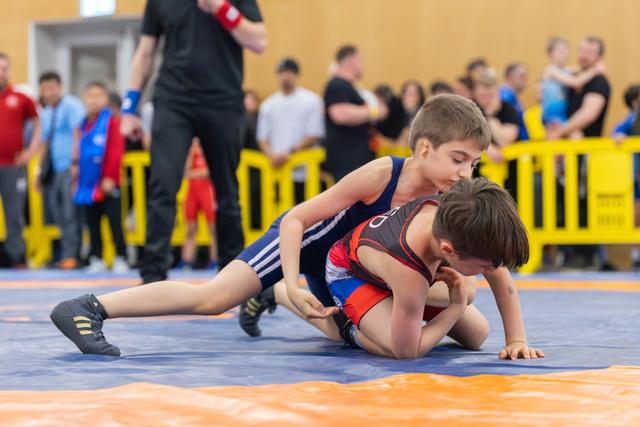 Two young wrestlers on a blue mat, one in navy pinning the other in red-black, intense focus during a youth bout.