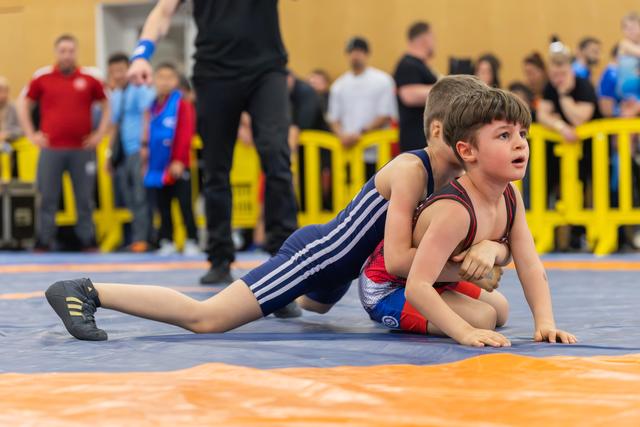 Two young boys wrestling on a blue mat, one in navy pinning the other in red, both showing intense concentration.