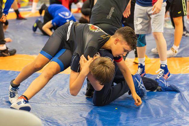 Two young wrestlers grapple on a blue mat, one boy dominating with focused intensity as he pins his opponent down.