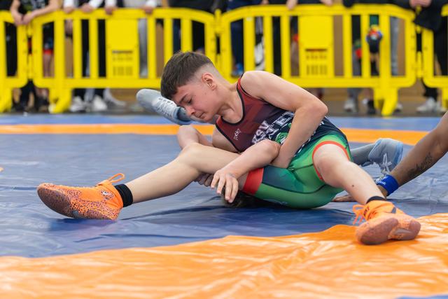 Determined young wrestler controls opponent on blue mat, wearing vibrant orange shoes and red-green singlet.