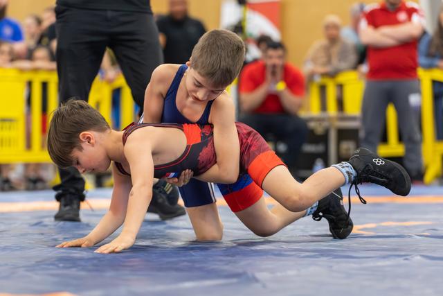 Two young boys wrestle intensely on a blue mat, one in blue dominating over the other in red, at an indoor sports event.