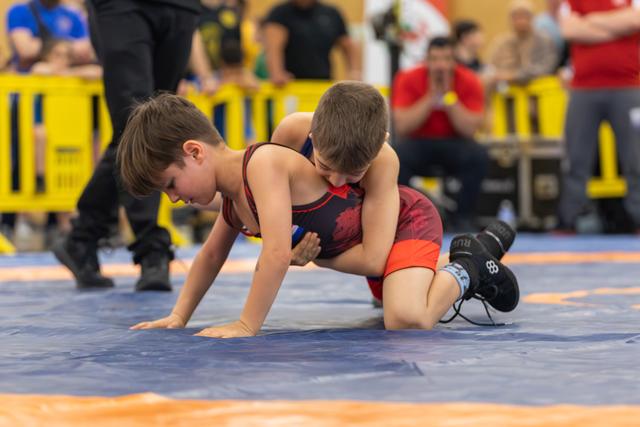 Two young boys wrestling intensely on a blue mat, one applying pressure from above while the other braces on all fours.