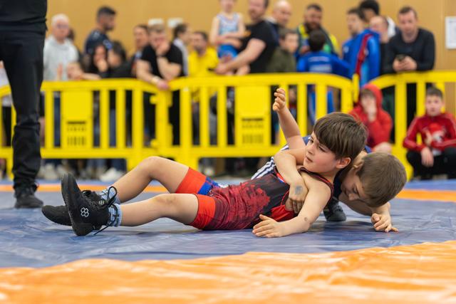 Two young boys wrestling on a blue mat, one attempting a pin while the other raises his arm, crowd watching behind barriers.
