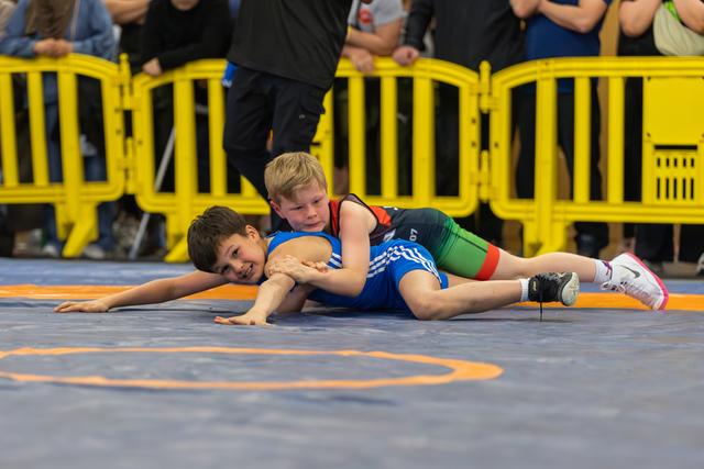 Boy in blue singlet pinned on mat, smiling, as opponent in black and green holds him down at indoor youth match.