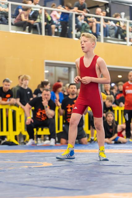 Young wrestler in red Adidas singlet stands focused on the blue mat, crowd watching from the stands behind.