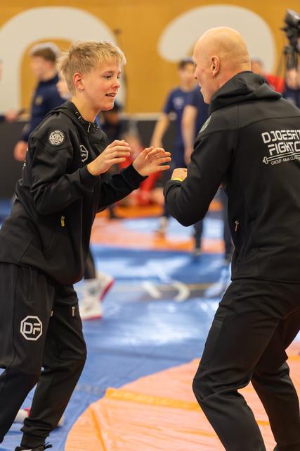 A young blond boy practices striking technique with a bald coach in black tracksuits on colorful gym mats.
