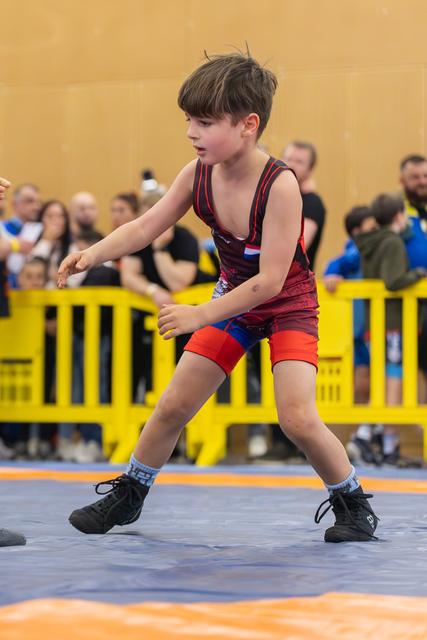 Young boy in red wrestling singlet crouches in a defensive stance on the mat, focused and alert during a match.