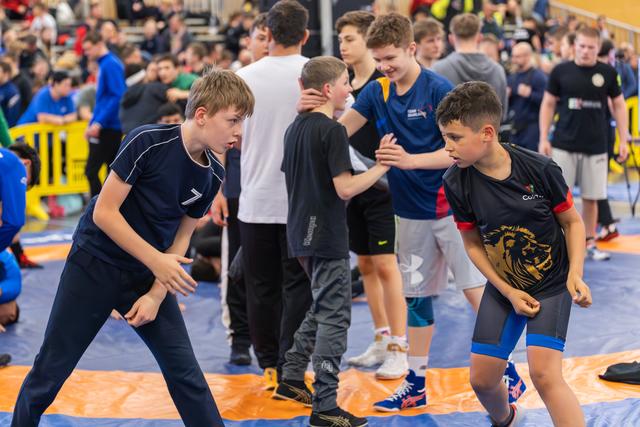 Two young wrestlers face off on the mat, both crouched in focused, determined stances during a busy indoor sports event.