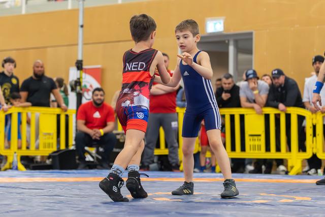 Two young boys in singlets square off on a blue mat, one wearing NED colors, at an indoor youth wrestling event.