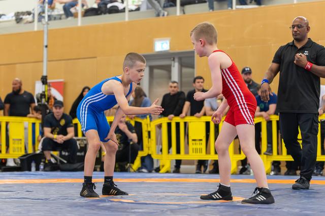 Two young wrestlers in blue and red singlets face off on a mat, referee watching closely in an indoor sports hall.