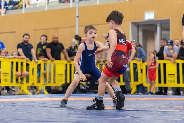 Two young boys in wrestling singlets face off on a blue mat, focused and poised, as spectators watch behind yellow barriers.
