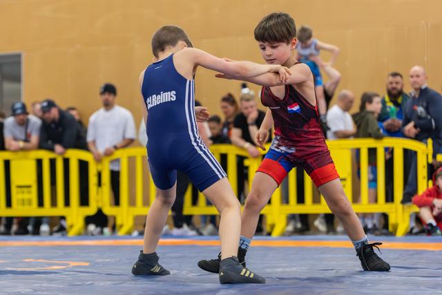 Two young wrestlers engage on the mat, one in blue 'Reims' singlet, the other in red Dutch colors, focused intensity.
