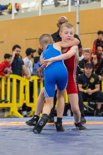 Two young boys locked in a grappling clinch, focused and determined, wrestling on a blue mat before a crowd.